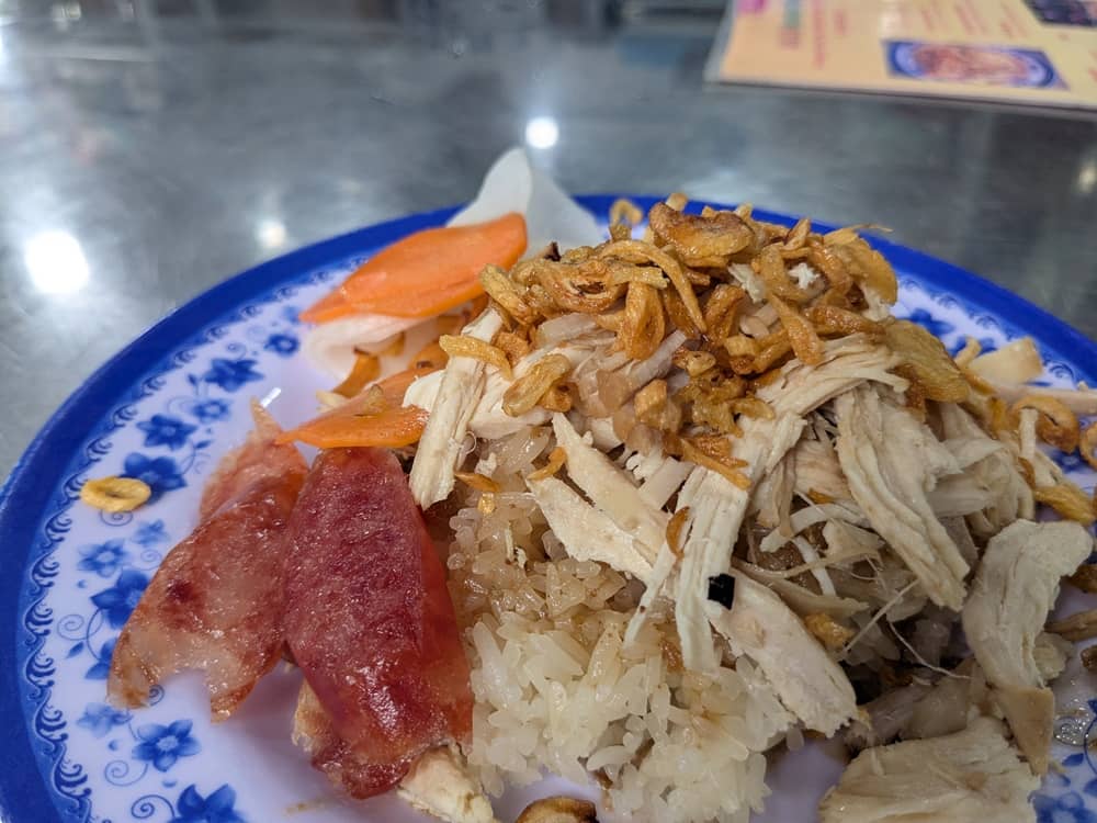 青い縁取りの皿に盛られた「中華ソーセージ入り鶏おこわ(Xôi gà lạp xưởng)」。しっとりした鶏肉に加え、赤みのある甘じょっぱい中華風ソーセージが添えられており、ボリューム感のある実食写真。