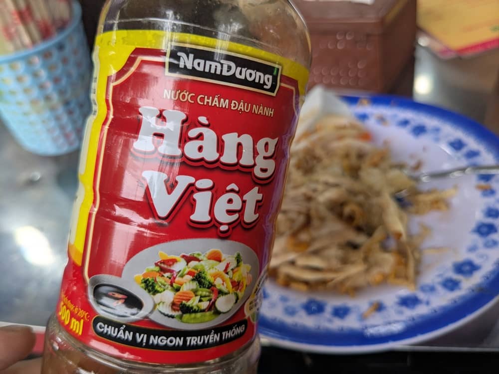 卓上のベトナム醤油(Nam Dương)。赤いラベルが特徴的なボトルの背景に、実食中の鶏おこわが写っており、ローカル店ならではの食事風景を伝えている。