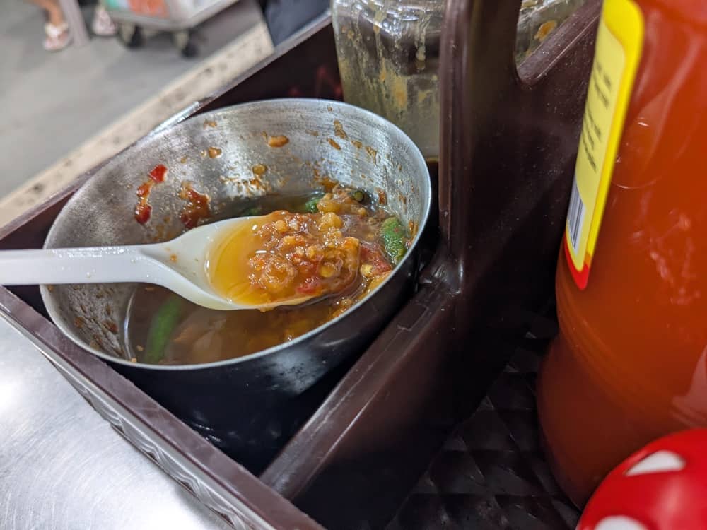 ソイガーナンバーワン特製の自家製チリソース。スプーンですくい上げたソースには刻んだニンニクや唐辛子がたっぷり入っており、食欲をそそる濃厚な色合いの調味料。