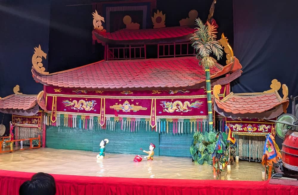 ロンヴァン水上人形劇のステージ。赤い屋根の豪華なセットを背景に、水面を泳ぐように動くコミカルな人形たち。