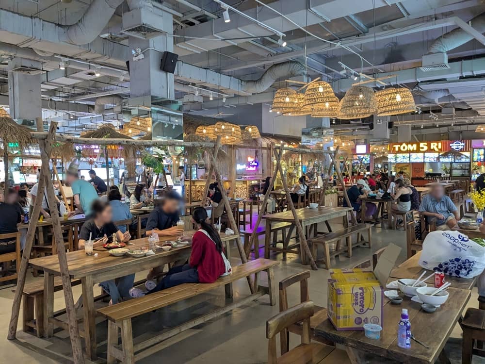 ホーチミン・TAKAプラザ(センスマーケット)内にある地下フードコートの様子。木製のテーブルとベンチが並び、多くの観光客や地元の人で賑わう。