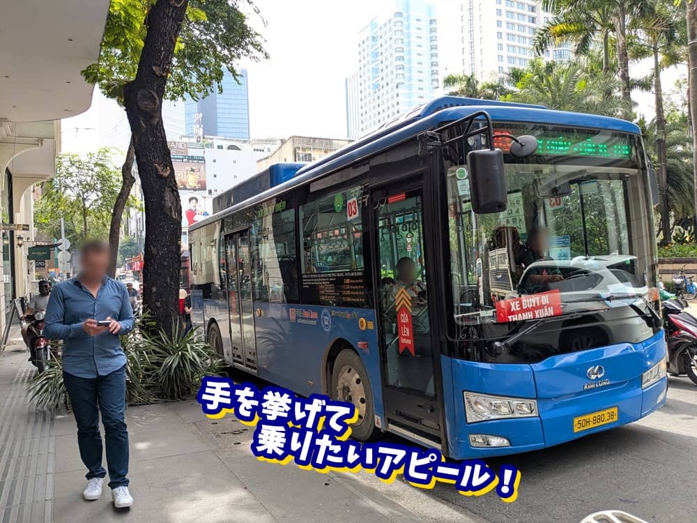 ベトナム・ホーチミンの街中を走る青い路線バス(03番)。バスの乗り方として、手を挙げて合図する必要があることを説明するテロップ付きの写真。